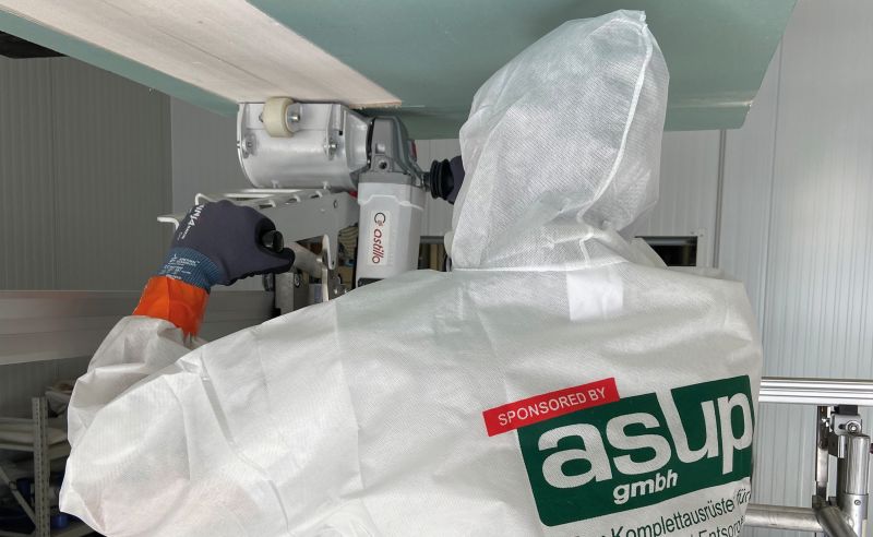 Eine Person in weißem Schutzanzug arbeitet an einer Maschine. Sie hält ein Werkzeug und ist mit einem technischen Gerät beschäftigt. Im Hintergrund sind industrielle Strukturen sichtbar, die auf eine technische Umgebung hinweisen.
